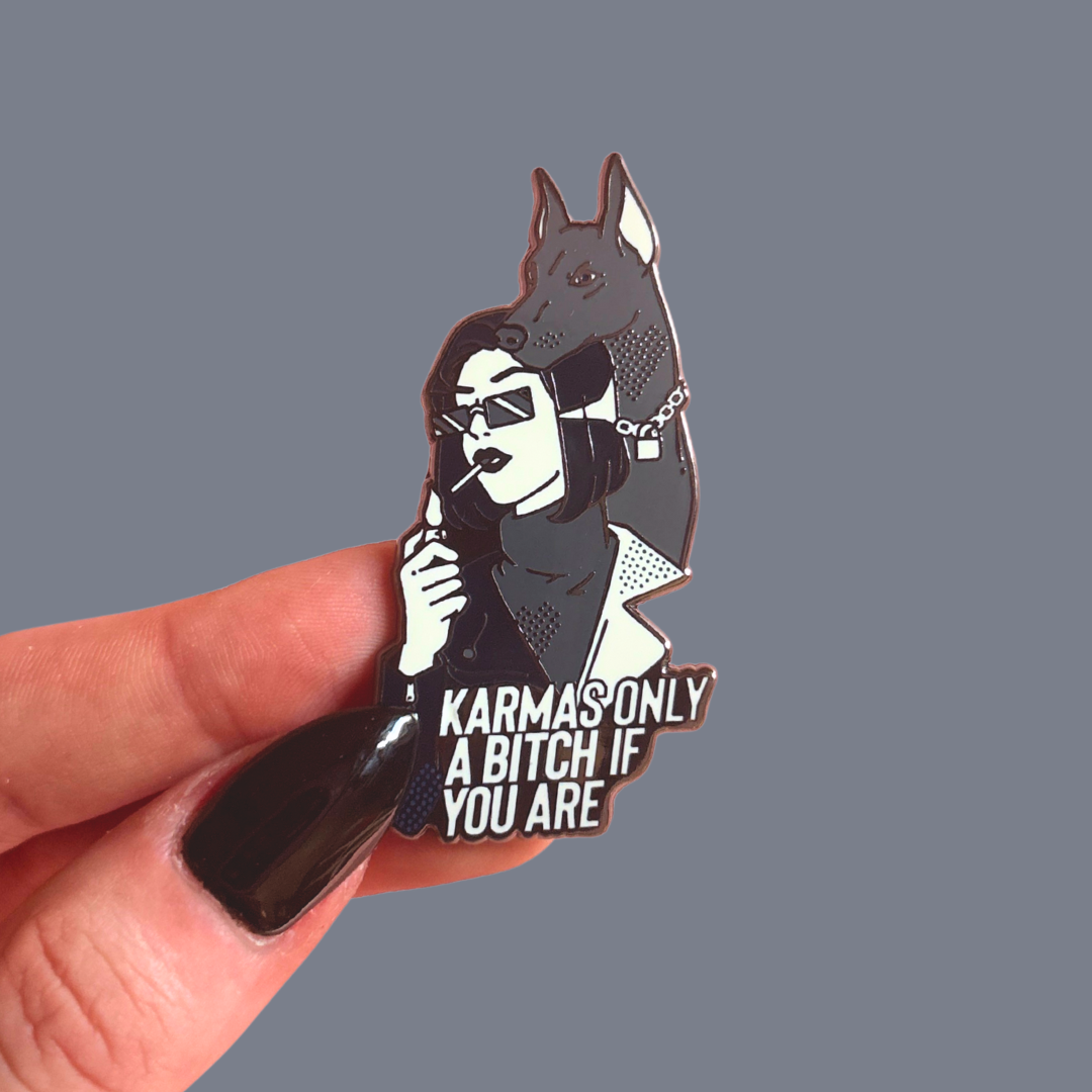 Pin with a design of a woman and dog, text 'Karmas only a bitch if you are', held by a hand against a gray background.