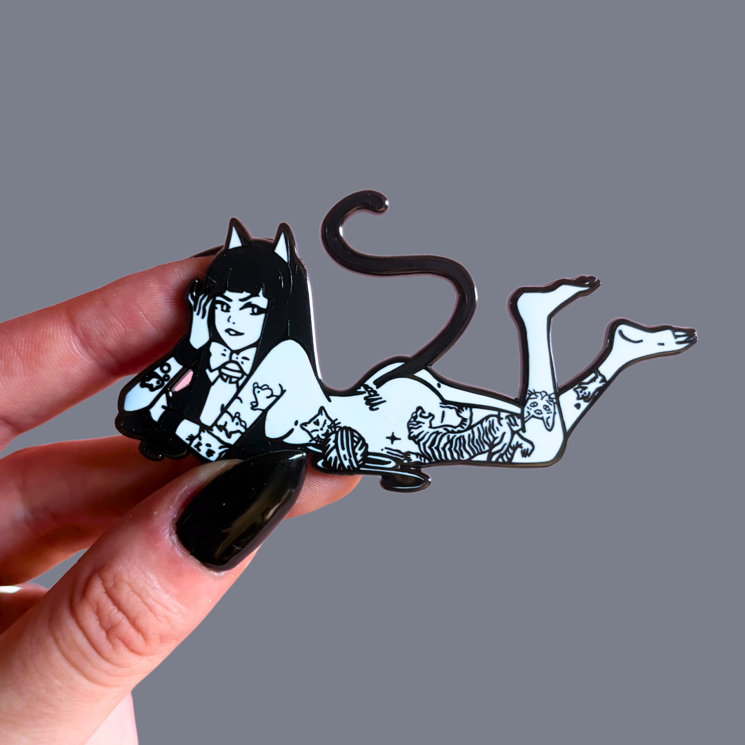Hand holding a black and white pin of a cat-like creature with a person's face.