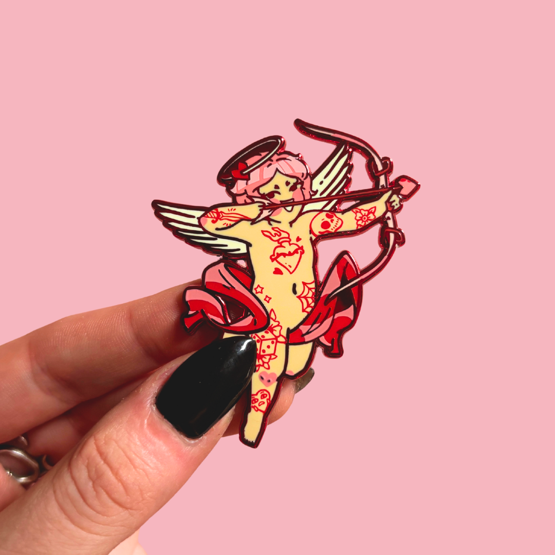 Pin with angel cherub design held by a hand against a pink background