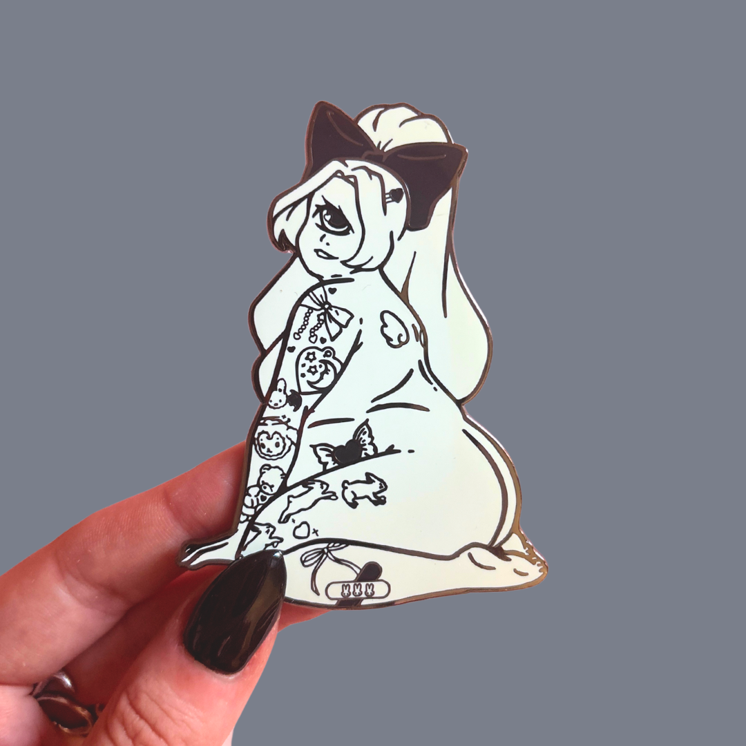 Pin of a cartoon cyclops girl character with tattoos held by a hand against a gray background
