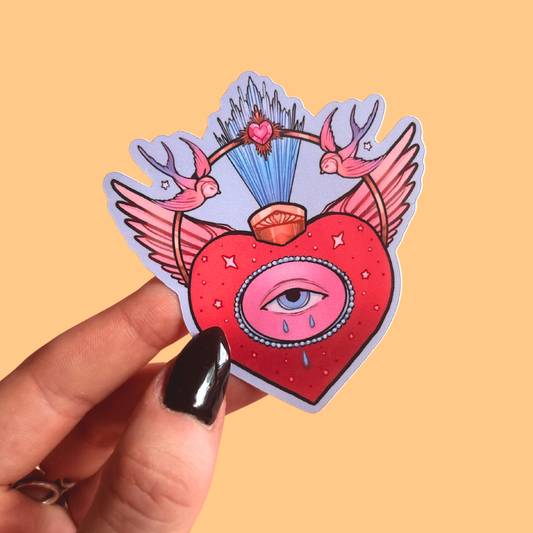 Heart-shaped sticker with sacred heart eye design held by a hand against a yellow background