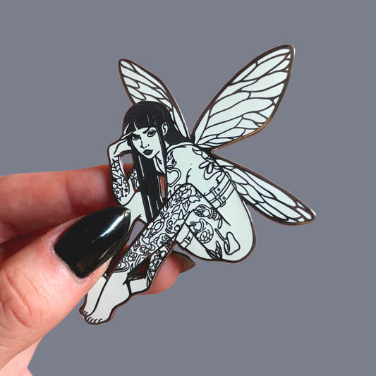 Tattooed fairy pin held by a hand against a gray background
