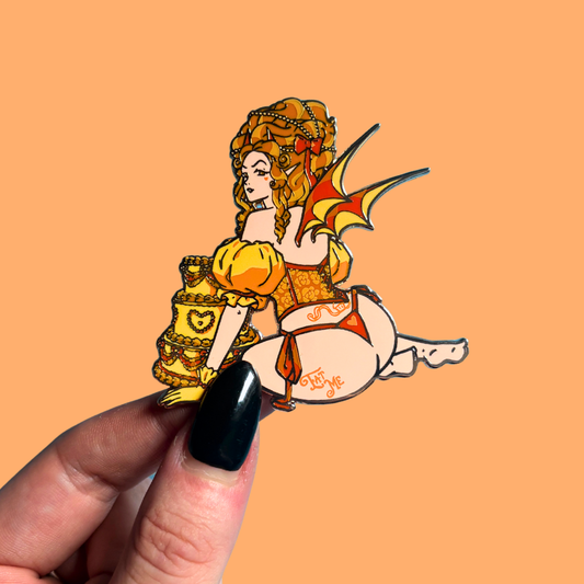 Hand holding a decorative pin of a fairy-like character and wings on an orange background.