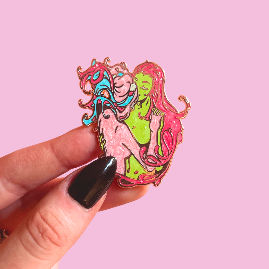 Colorful enamel pin of a 2 stylized characters held by a hand against a pink background