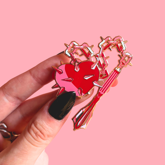 Heart-shaped pin with chain and spikes held by a hand against a pink background