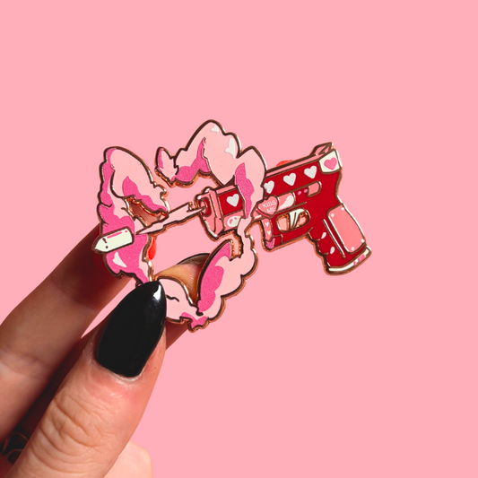 Pink and red pistol enamel pin with held by a hand against a pink background