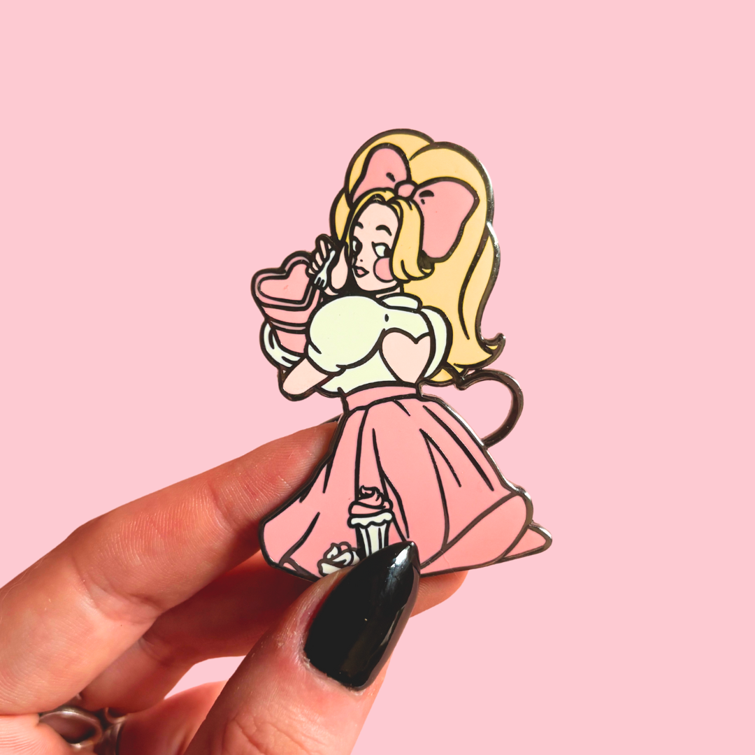 Cartoon character pin in a pink dress holding a cake, held by a hand with black nail polish on a pink background.