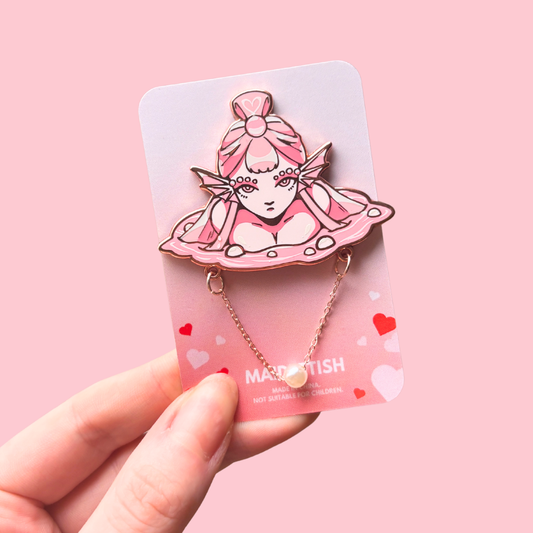 Pin with an illustrated character on a pink card held by a hand against a pink background