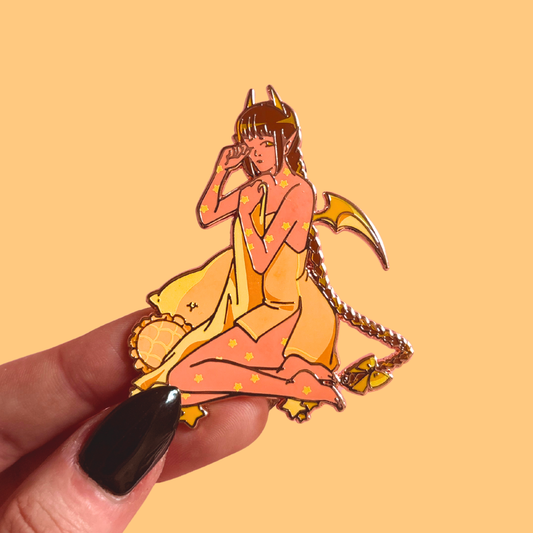 Hand holding a yellow enamel pin of demon character with a yellow and orange color scheme against a beige background
