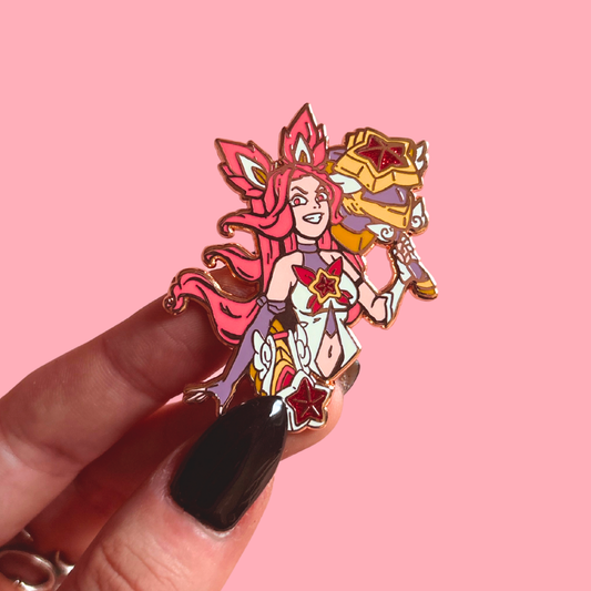 Enamel pin of a character with red hair and a crown held against a pink background