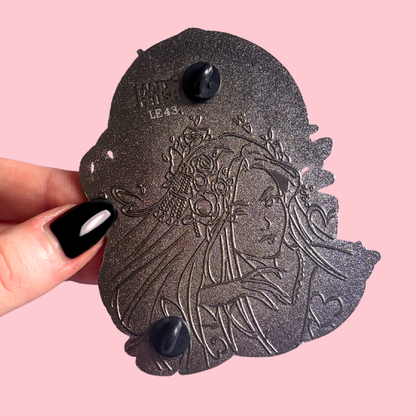 Decorative metal piece with engraved design held by a hand on a pink background