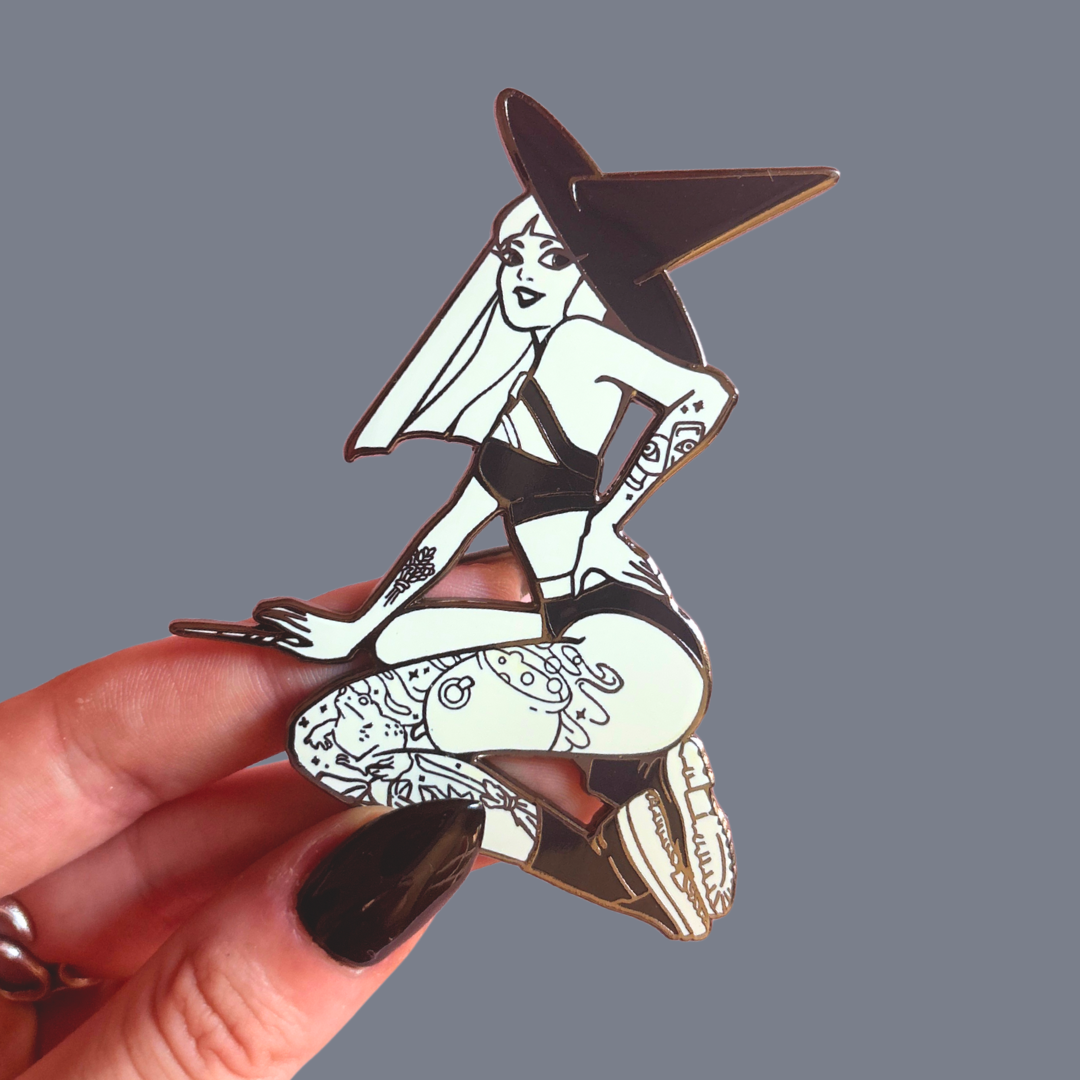 Pin with a stylized witch design held by a hand against a gray background