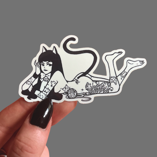 Sticker of a stylized cat-like figure held by a hand against a gray background