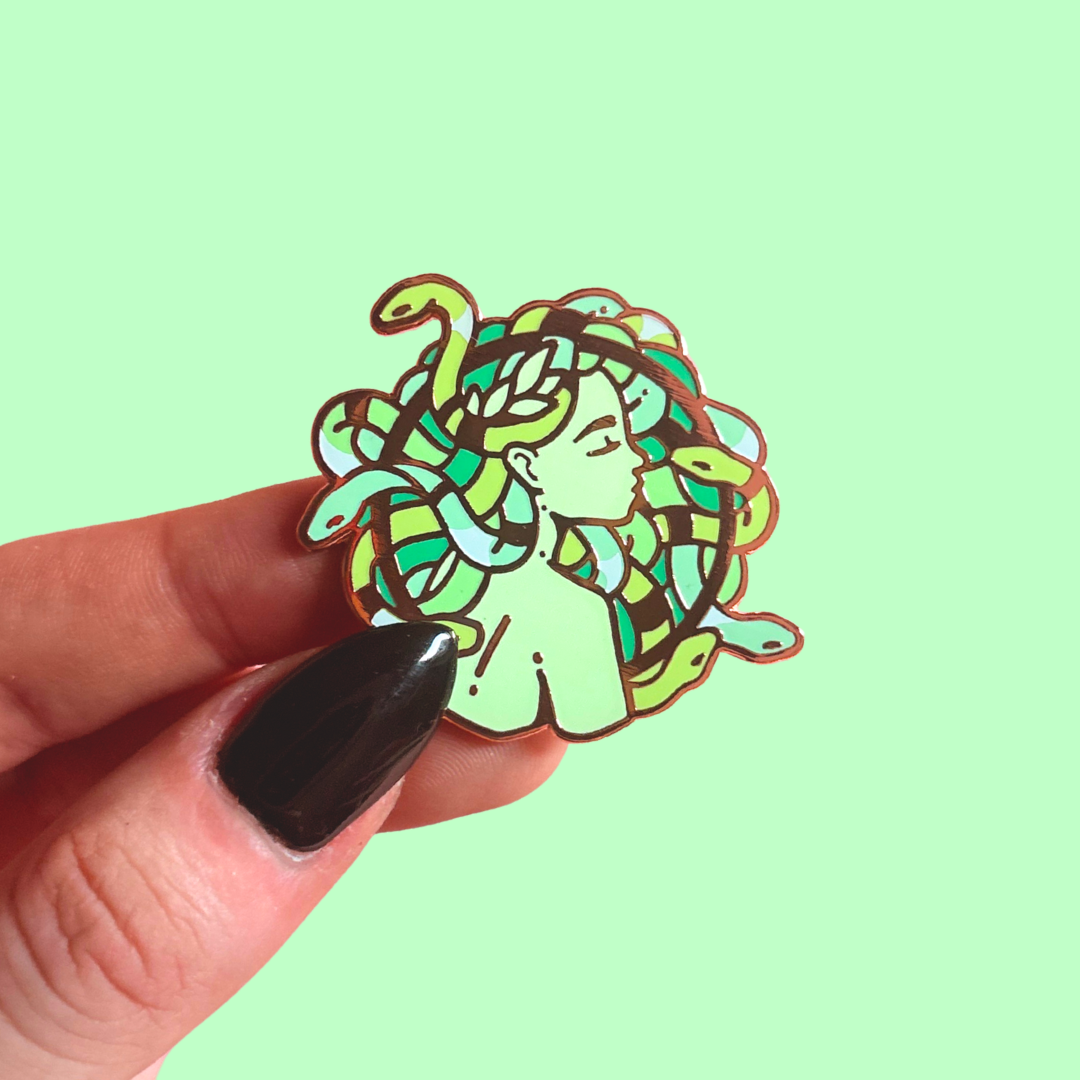 Medusa-themed pin held by a hand with black nail polish against a light green background